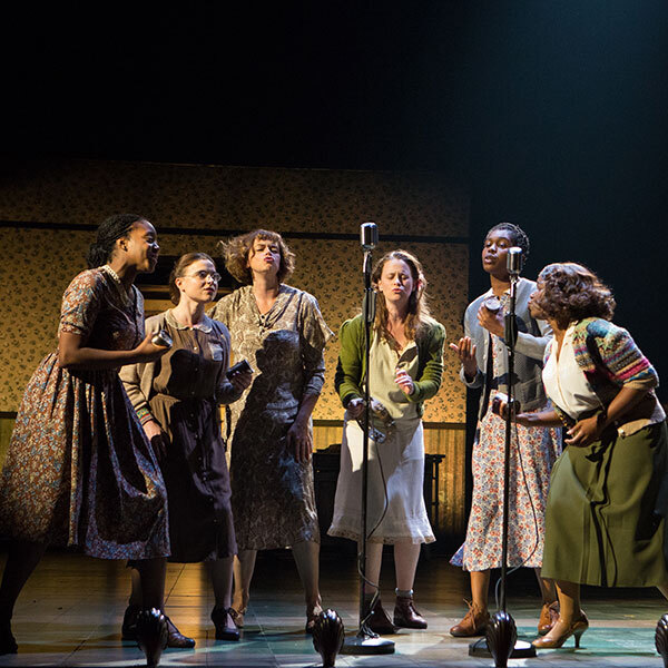 Women standing in a semi-circle singing into microphones.