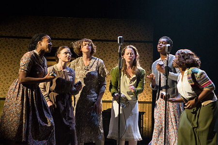 Women standing in a semi-circle singing into microphones.